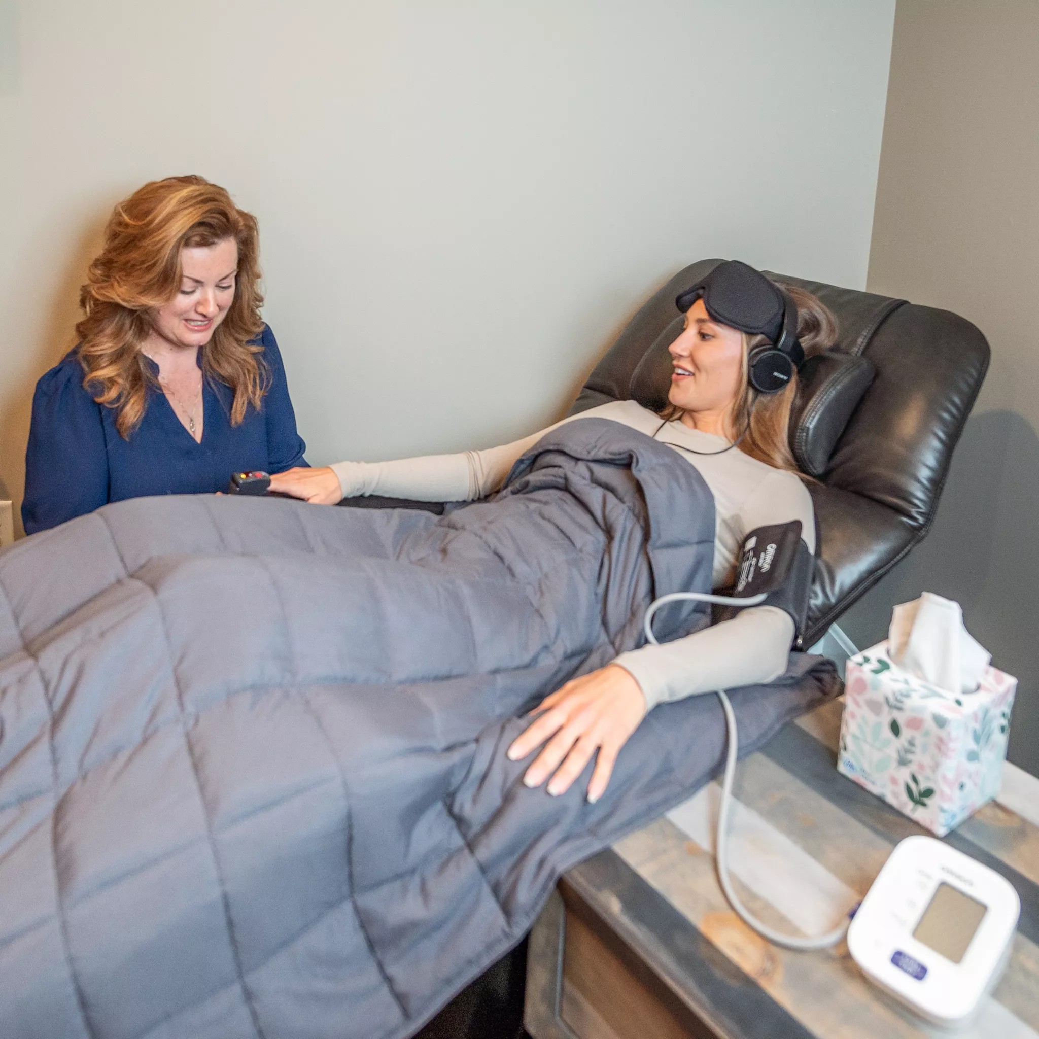 Dr. Monica Hurt seated beside a patient during a ketamine-assisted psychotherapy session, monitoring vitals while the patient rests comfortably with an eye mask and headphones
